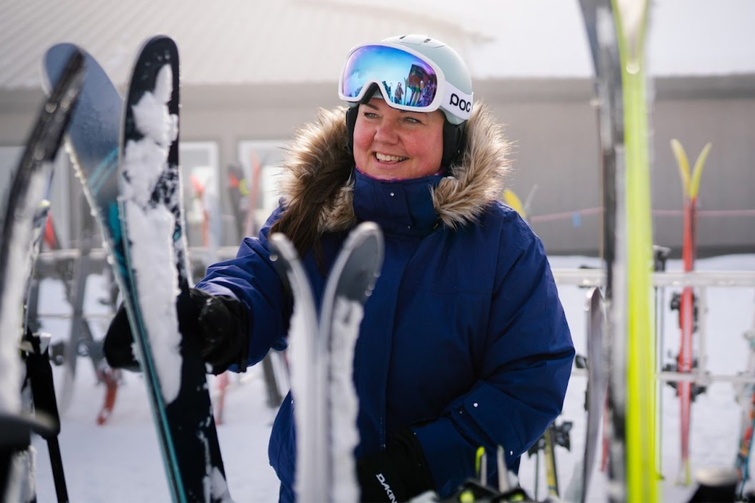 Plus size female skier wearing a white helmet and goggles reaching for skis in a ski rack in the snow.