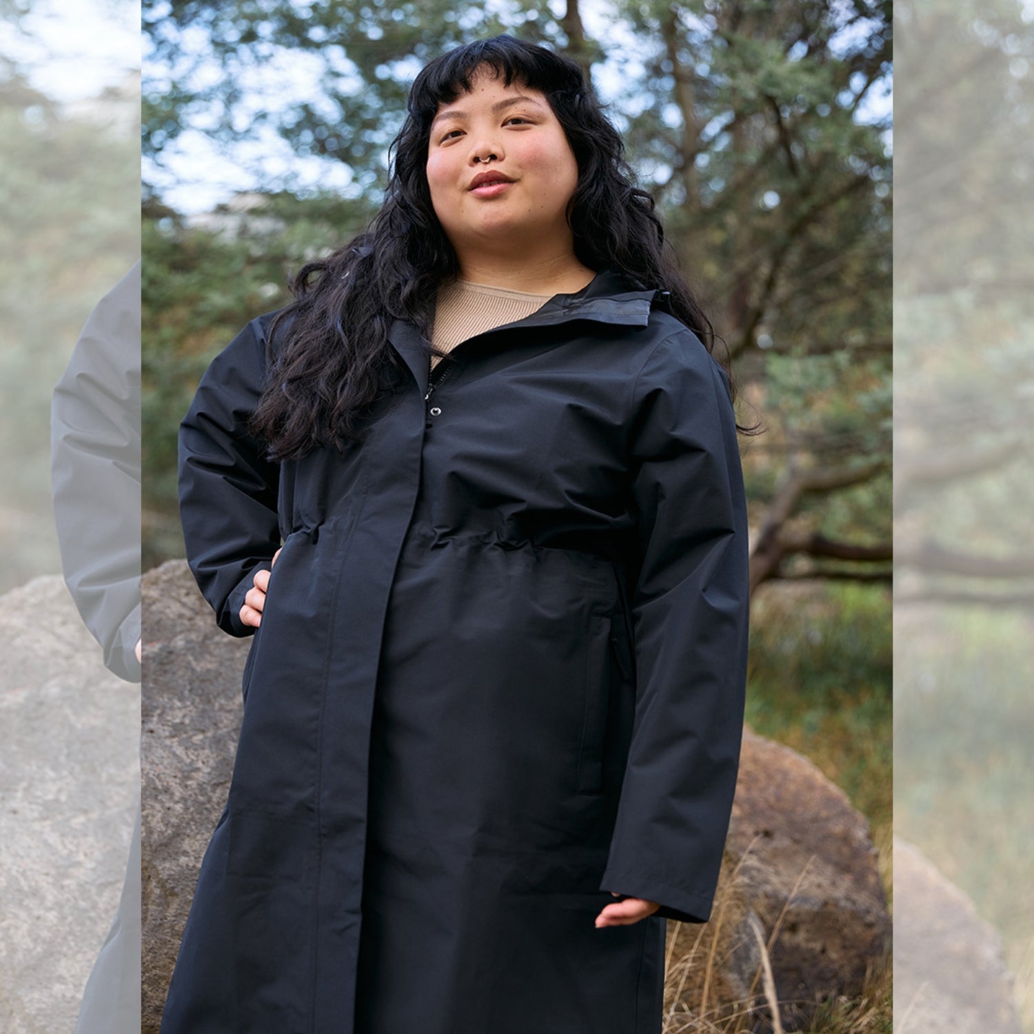 A woman standing outdoors wearing an Amble Outdoors waterproof and breathable long black rain jacket with a large hood, snap buttons, and a YKK zippered front.
