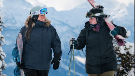 Two plus-size friends talking on a snowy mountain summit, with one wearing the Cartel Brooklyn Stretch Women's Plus Size Ski Jacket in dark blue while carrying a snowboard.