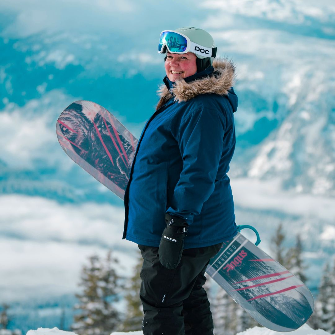 Smiling woman holding a snowboard on a mountain summit, wearing the Cartel Brooklyn Stretch Women's Plus Size Ski Jacket in blue.