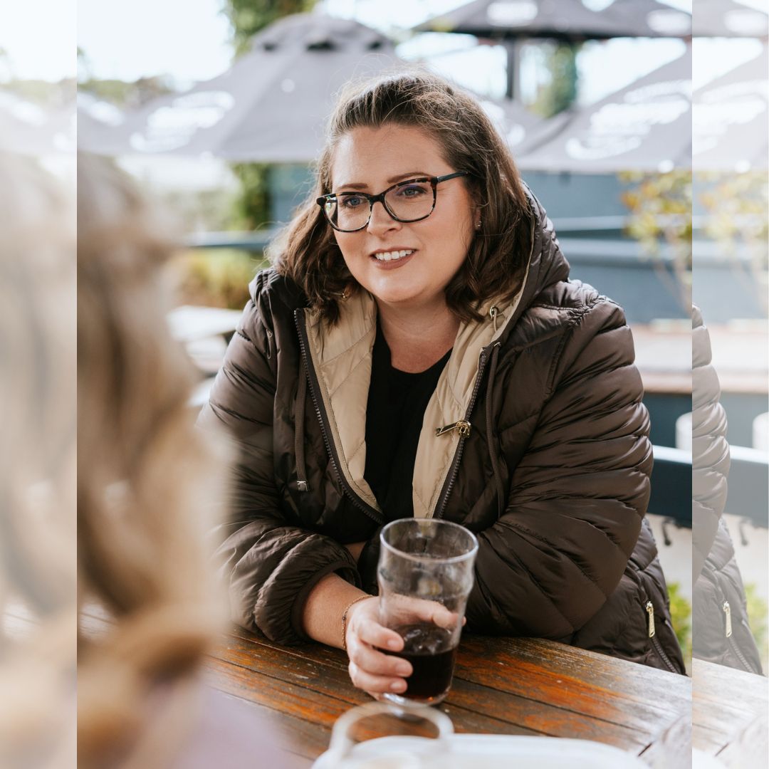 Woman with glasses sitting at an outdoor patio table wearing the unzipped chocolate brown Moke Arnie Women's Reversible Puffer Coat.