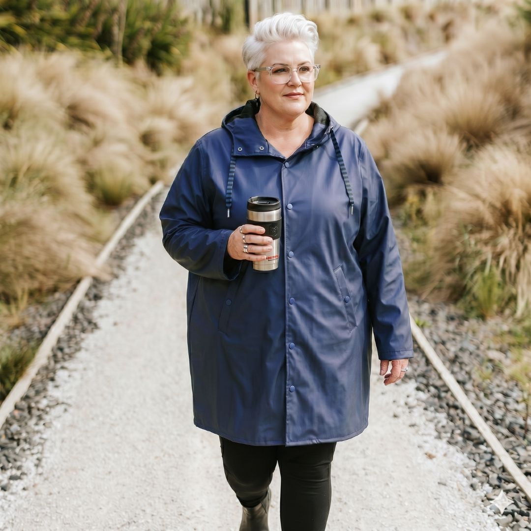 Front view of a smiling woman with glasses walking outdoors in the buttoned metallic blue Moke Charlie Women's Rain Coat.