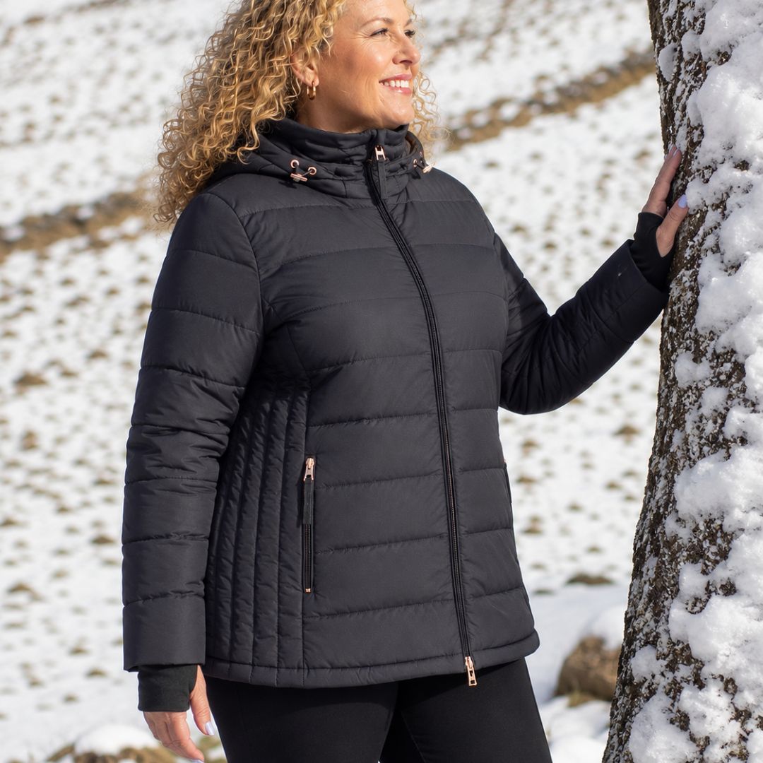 Side view of a smiling blonde woman leaning against a snow-covered tree wearing the black Moke Luciana Women's Down Puffer Jacket.