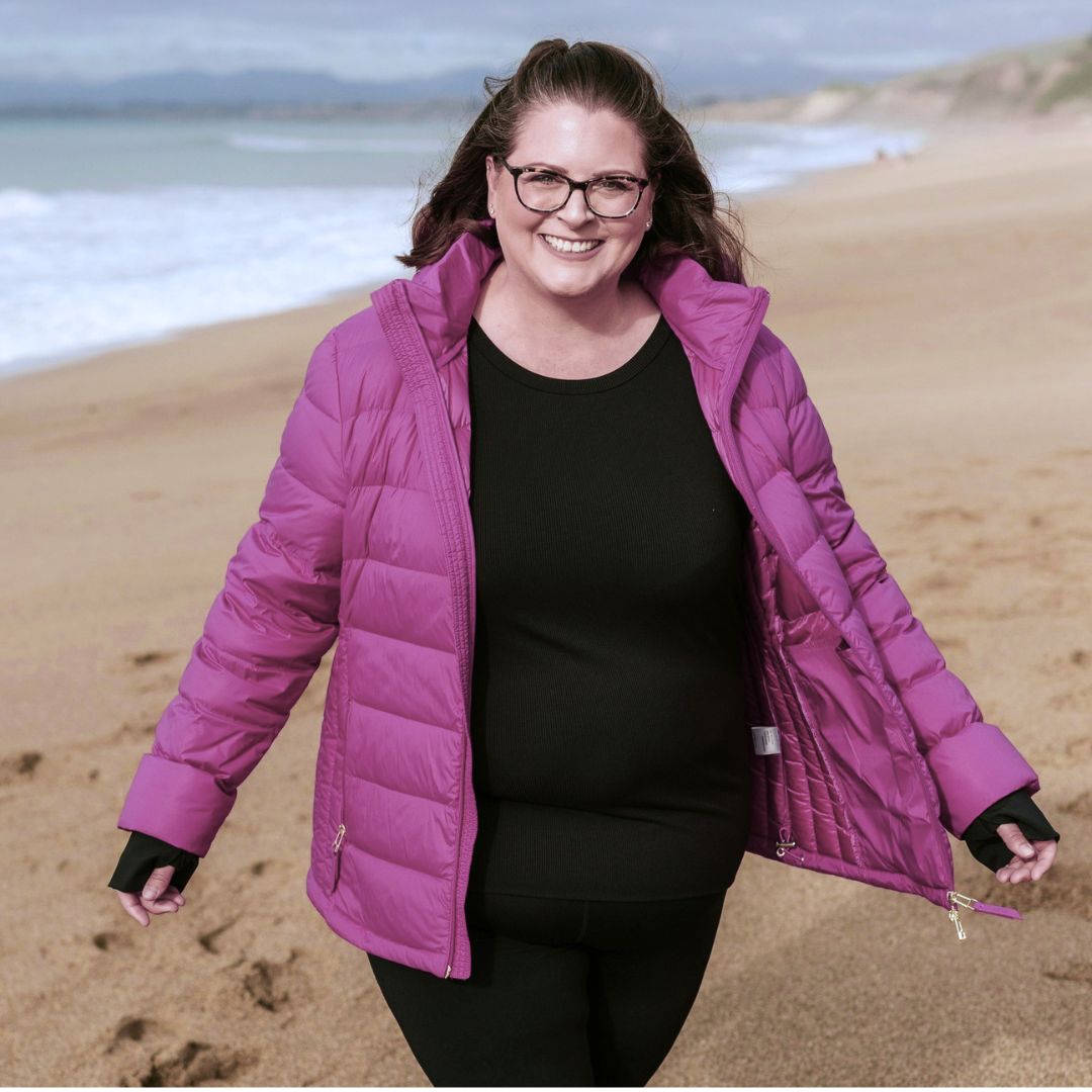 Smiling woman walking on a sandy beach wearing an unzipped Moke Luciana Women's Down Puffer Jacket in rose violet.