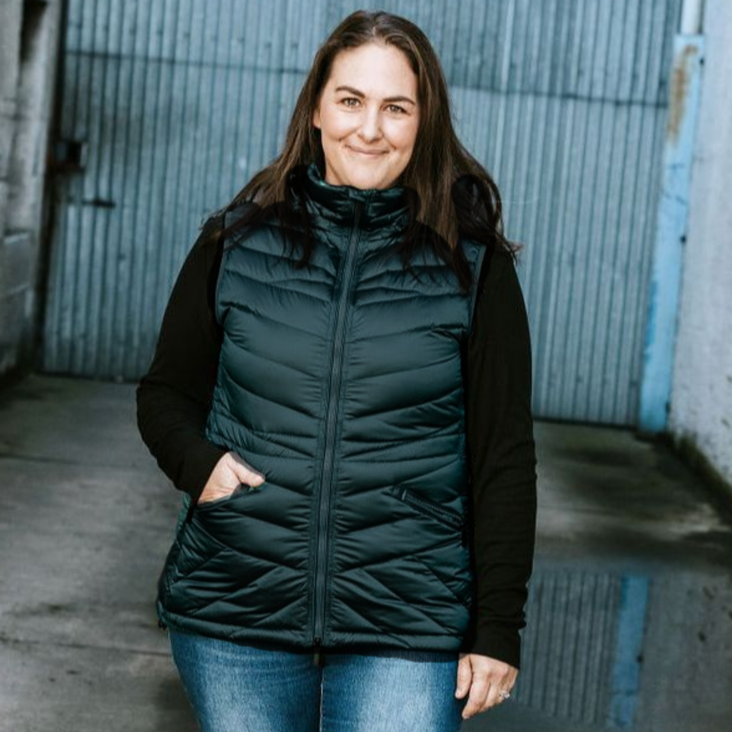 Curvy woman wearing a Moke black down puffer vest standing in front of a metal gate. Modelling plus size clothing as sold on Plus Outdoor