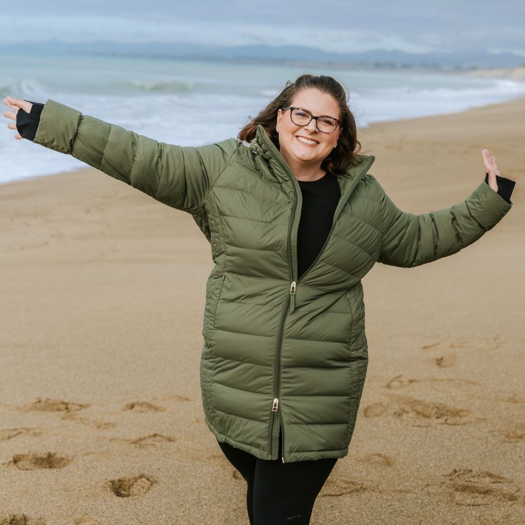 Laughing woman with arms outstretched walking on a sandy beach wearing the Moke Sydney Packable Down Coat in olive green.