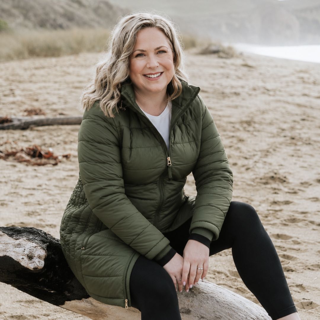 Smiling blonde woman sitting on a log at the beach wearing the olive green Moke Sydney Packable Down Coat.