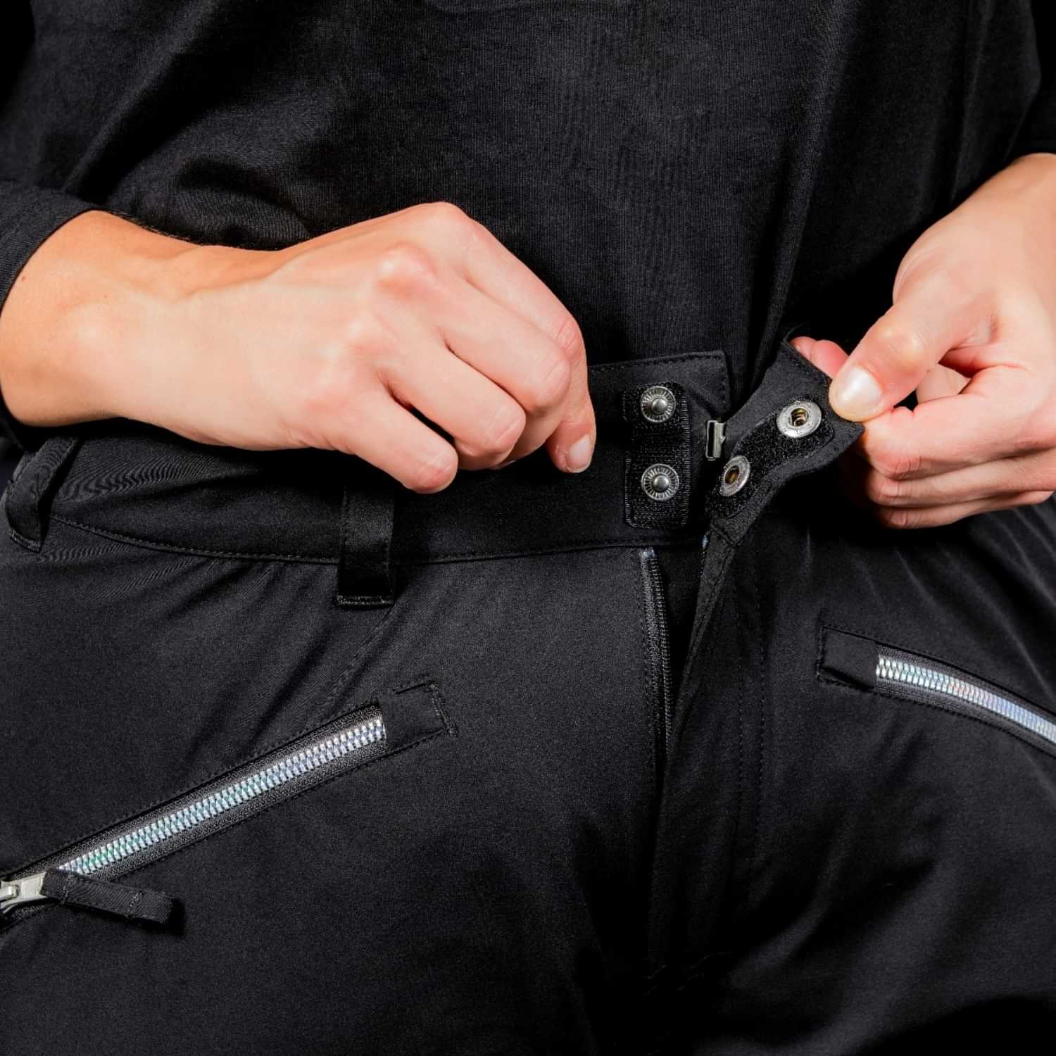 Close-up view of hands fastening the double snap-button and hook waist closure of Nobody's Princess Zali Plus Size Snow Pants in black.