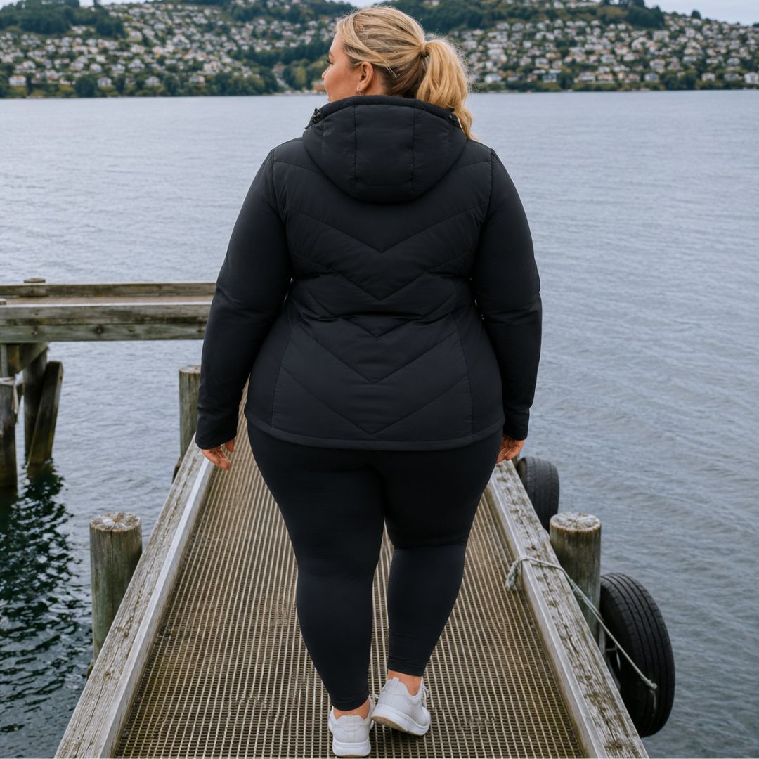 Rear view of a woman with a ponytail walking on a wooden pier, showcasing the chevron baffling on the back of the black Olive Packable Down Jacket.
