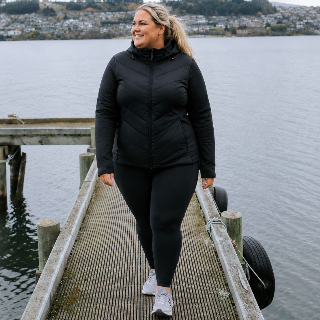 Full-body front view of a smiling woman walking down a pier over the water wearing the black Olive Packable Down Jacket with black leggings.