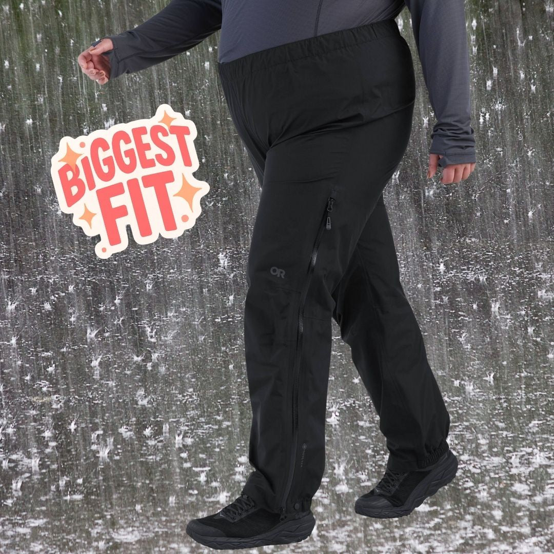 A plus-size person wearing black outdoor research rain pants and black shoes, shown from the shoulders down against a snowy, rain-slicked ground. A graphic badge on the left reads 'BIGGEST FIT'.