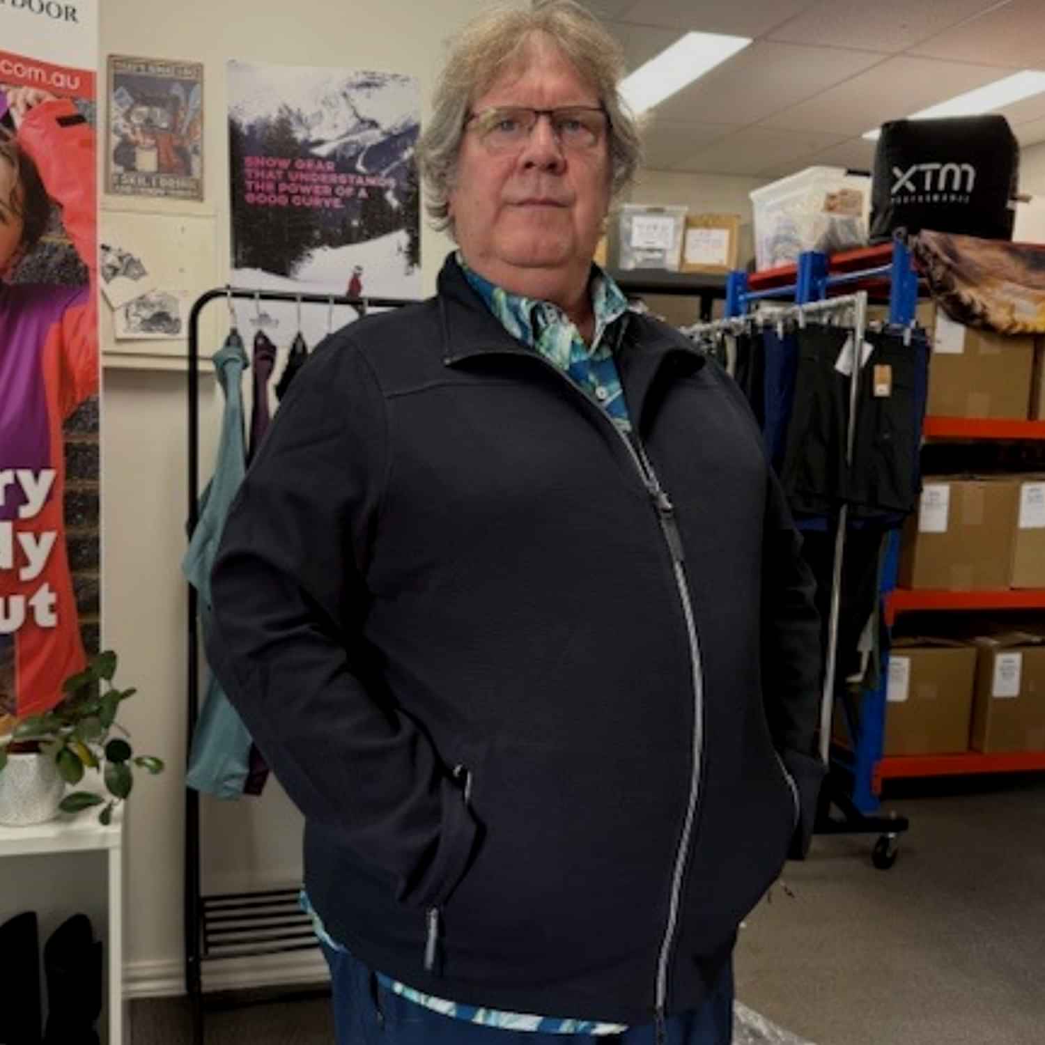 Three-quarter view of a man standing with his hands in his pockets, wearing a black Stoy Powerstretch Men's Plus Size Zipped Jacket inside a retail store.