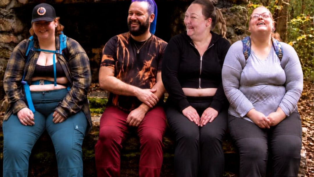 Four plus-size people sitting together outdoors in front of a stone fireplace or log cabin entrance, with trees and fallen leaves visible in the background.