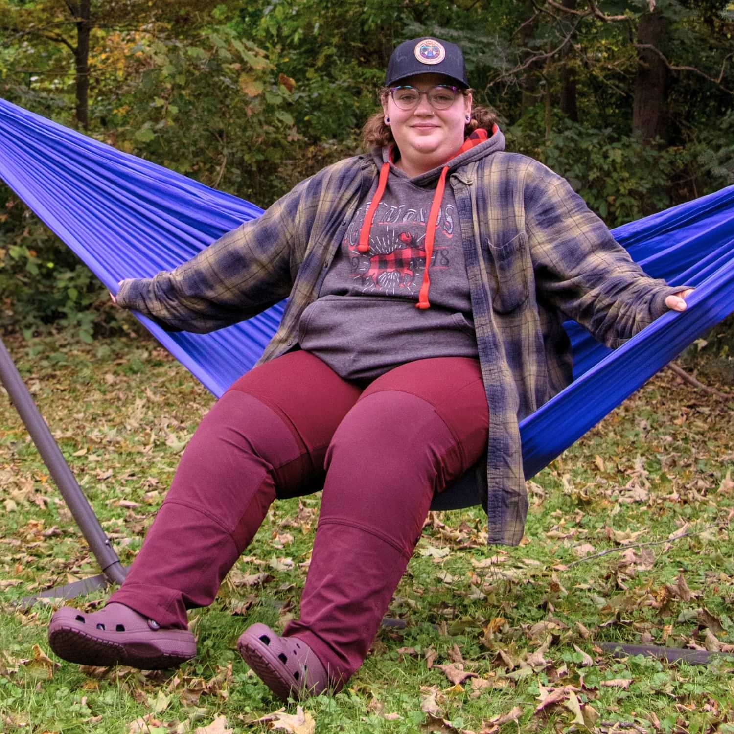 Plus size model relaxing in a blue hammock outdoors wearing Thicket Adventure Brecken Travel Pants BELLY FIT in berry red, styled with a hoodie and flannel for camping and hiking comfort.