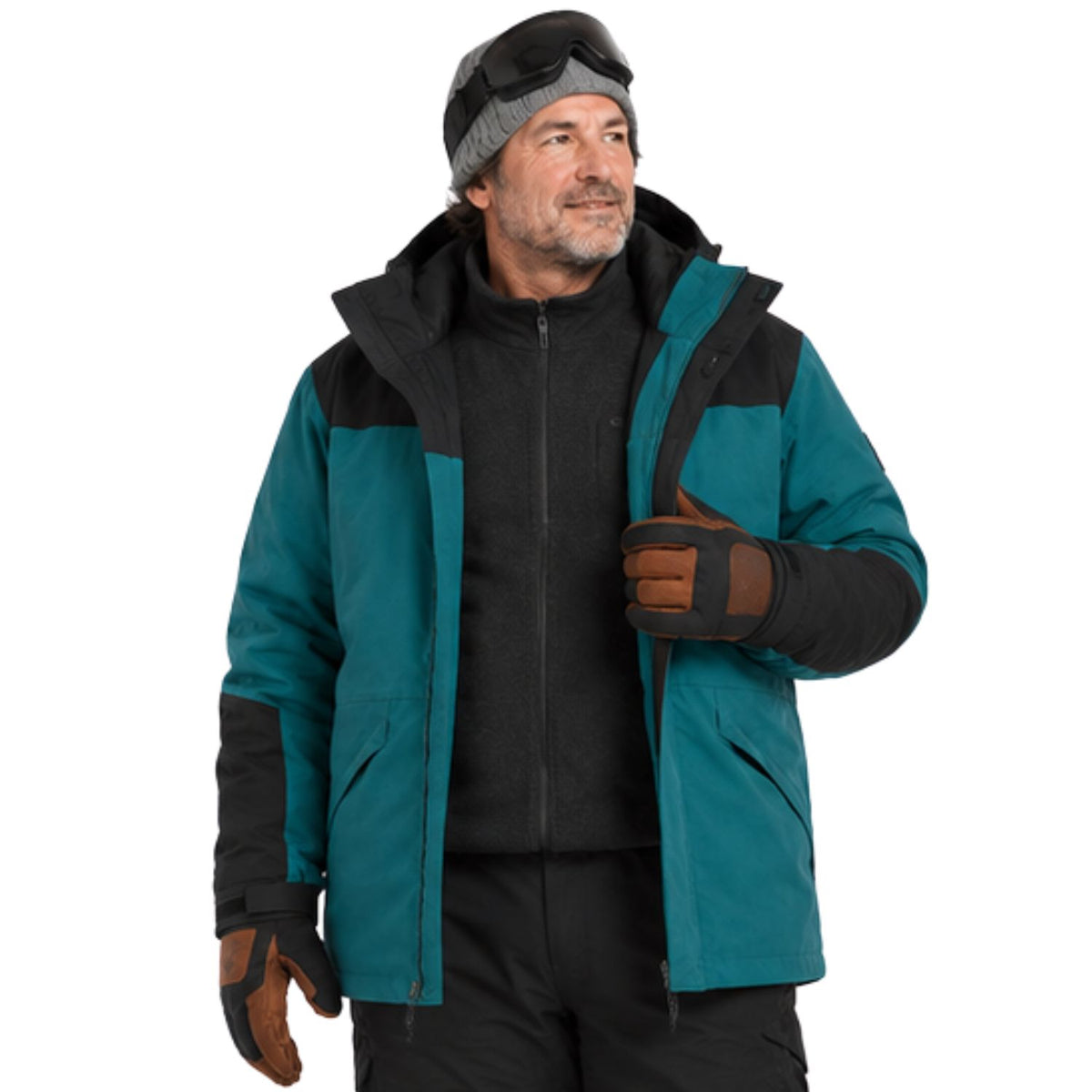 A happy skier man wearing a teal and black ski jacket, black fleece, brown gloves, a gray beanie, and ski goggles, standing against a solid white background.