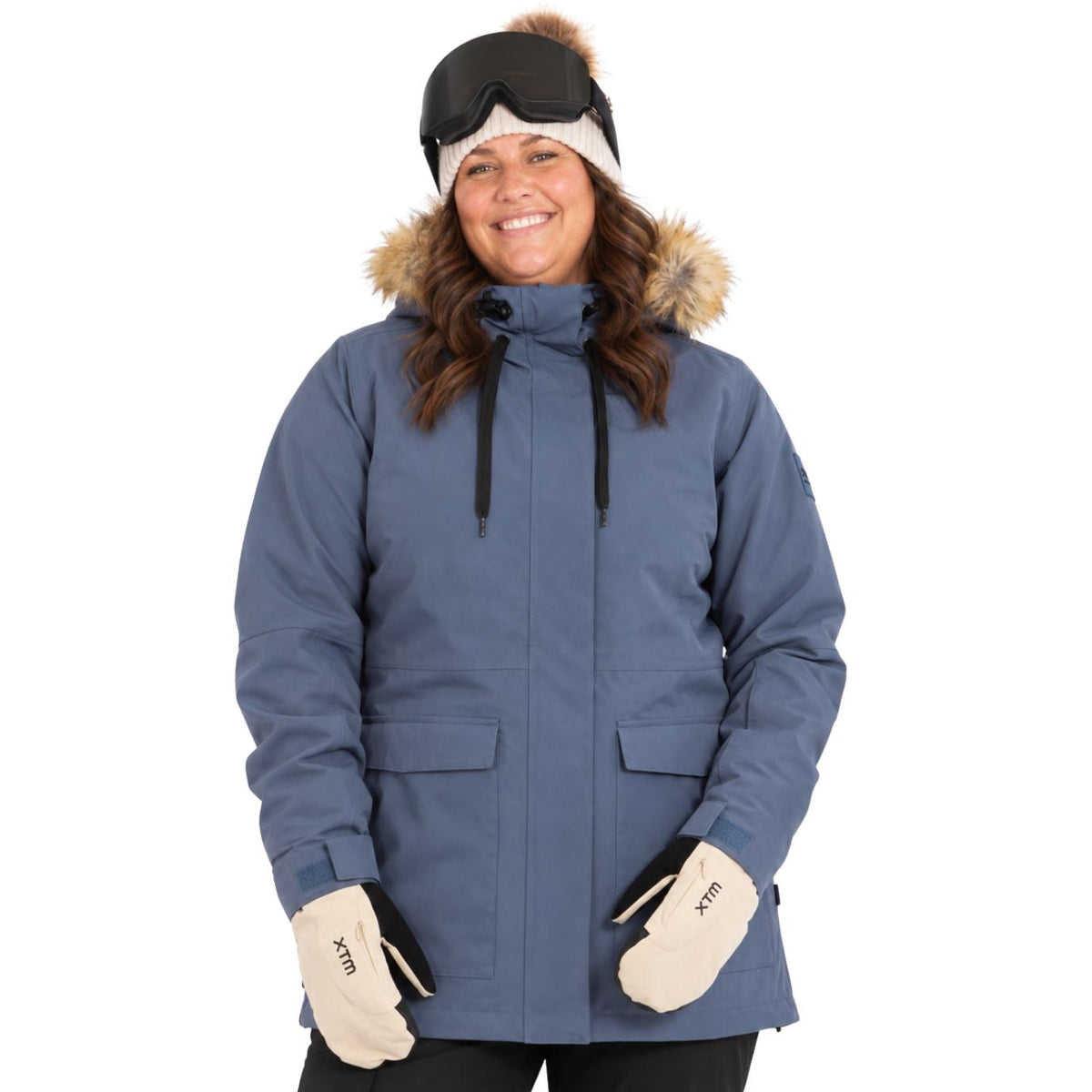 A size 20 woman wearing a slate blue ski jacket with a brown fur-trimmed hood, black goggles, beige gloves, and black pants, photographed against a solid white background.