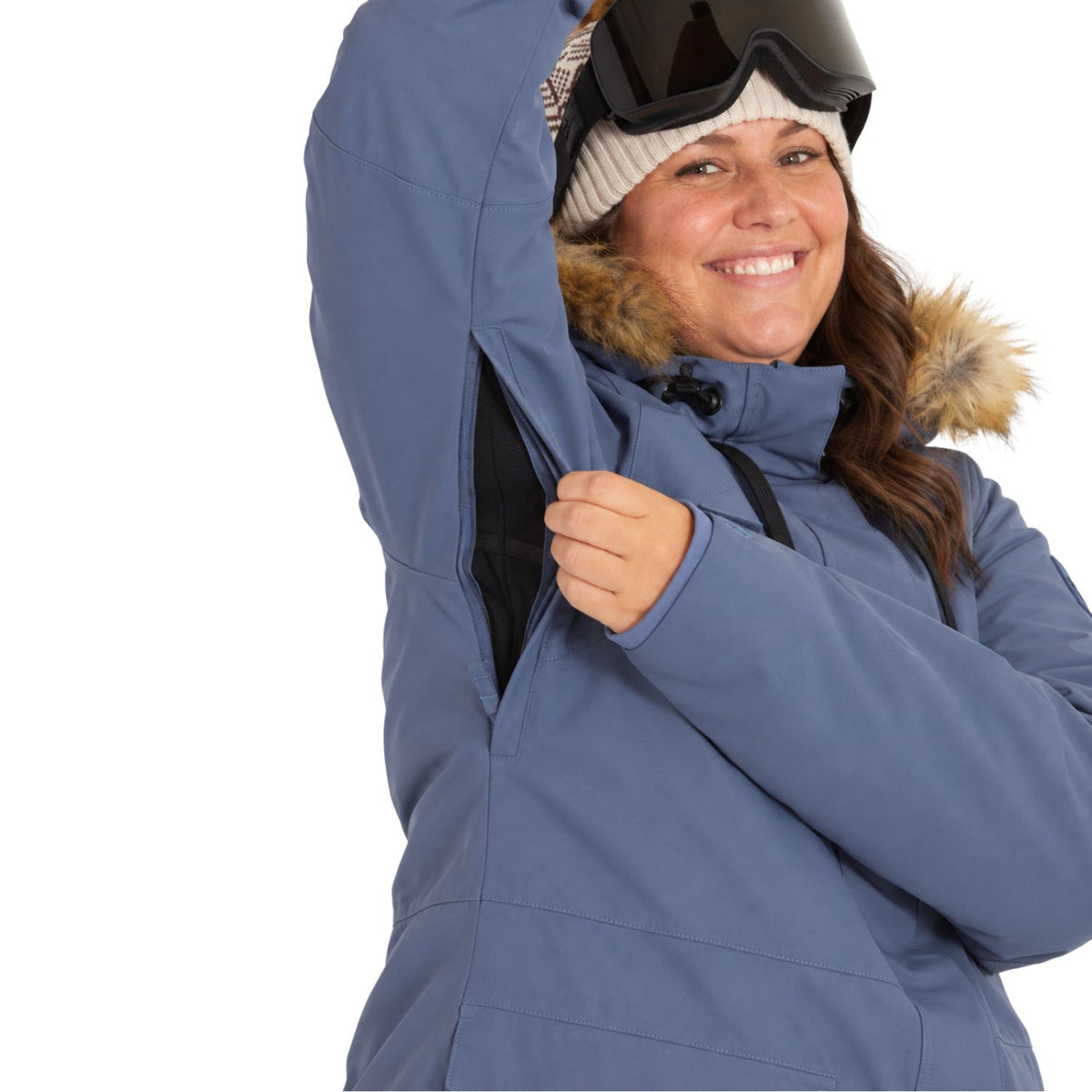 A woman wearing a slate blue ski jacket with a fur-trimmed hood, black goggles, and a cream knit beanie, smiling while demonstrating a zippered pocket on her sleeve against a white background.