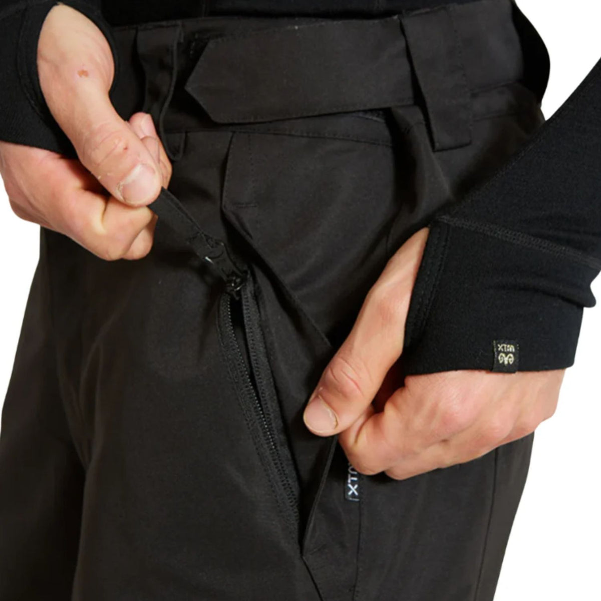 Close-up of a person wearing black technical pants, opening a zippered pocket with both hands. A small black G100 label is visible on the cuff of the right sleeve.