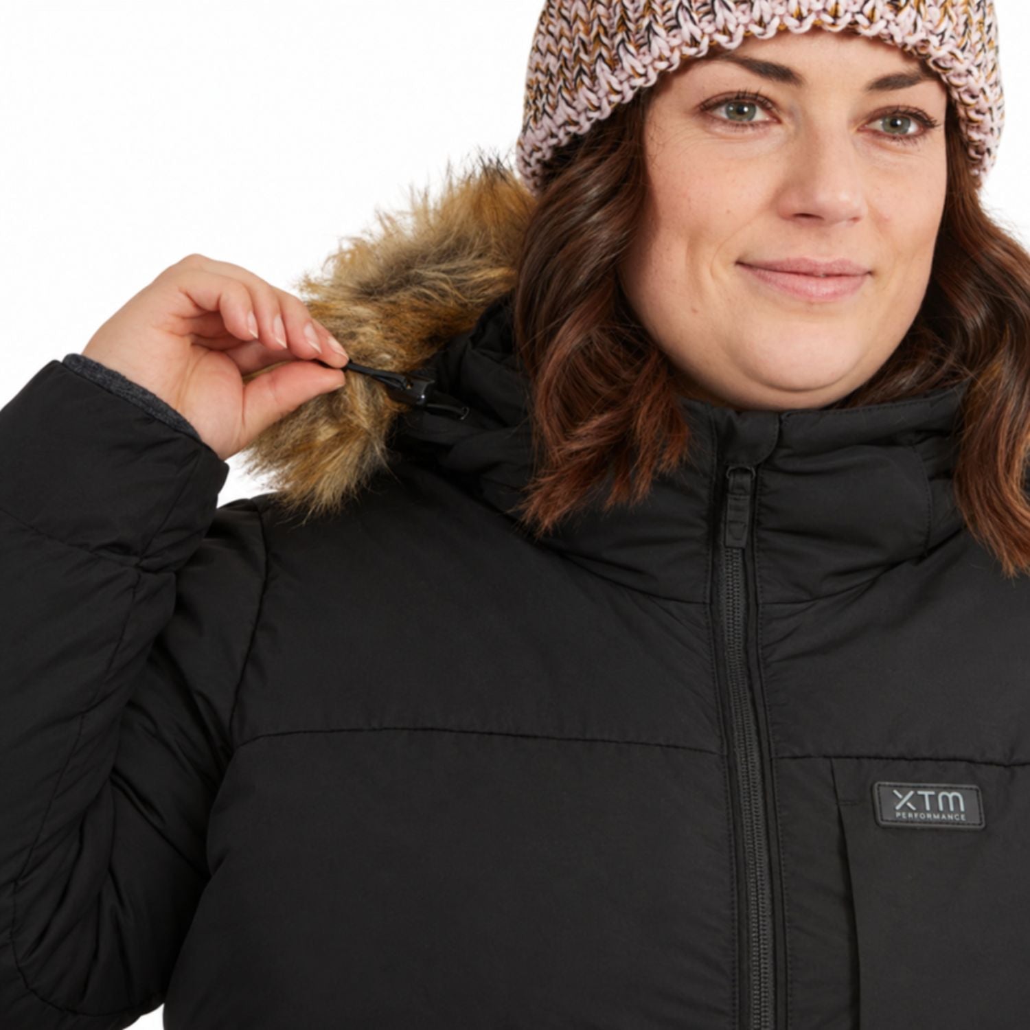 A close-up of a size 22 woman wearing a black winter puffer jacket with a brown faux-fur trimmed hood and a knit beanie. The jacket has a visible 'XTm' logo patch on the chest.