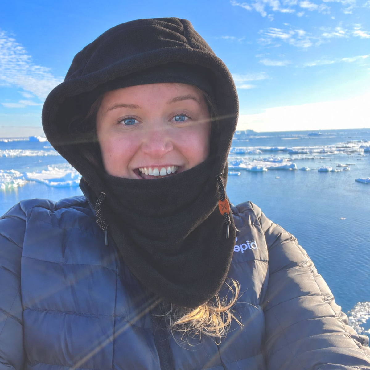 A plus size woman wearing a black fleece hooded balaclava face covering and a gray puffer jacket, smiling outdoors with icy water and ice floes in the background.