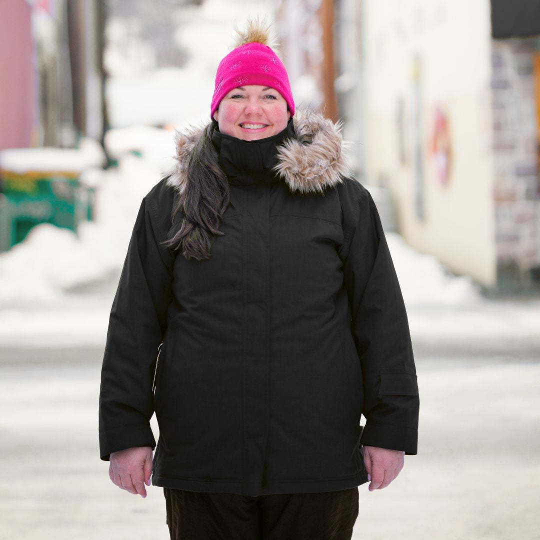 Smiling woman wearing the Cartel Brooklyn Stretch Women's Plus Size Ski Jacket in black with a pink beanie and a faux-fur trimmed hood.