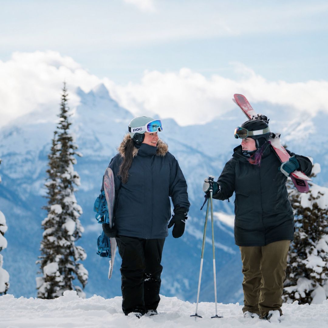 Two friends talking on a snowy mountain summit, with one wearing the Cartel Brooklyn Stretch Women's Plus Size Ski Jacket in dark blue while carrying a snowboard.