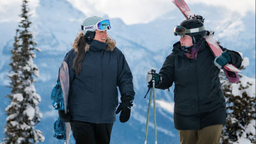 Two plus-size friends talking on a snowy mountain summit, with one wearing the Cartel Brooklyn Stretch Women's Plus Size Ski Jacket in dark blue while carrying a snowboard.
