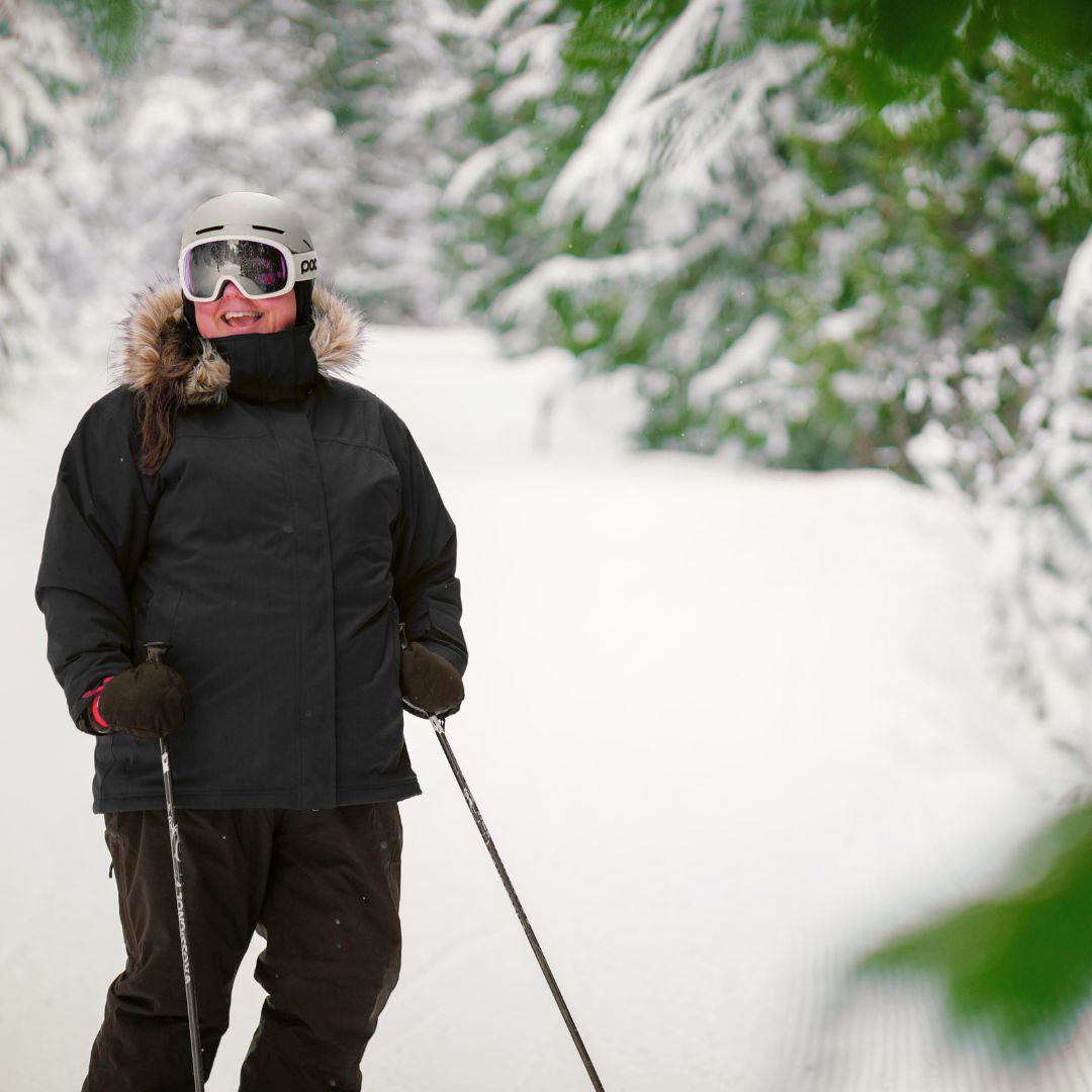 Woman skiing down a snowy forest trail wearing the Cartel Brooklyn Stretch Women's Plus Size Ski Jacket in black with a helmet and goggles.