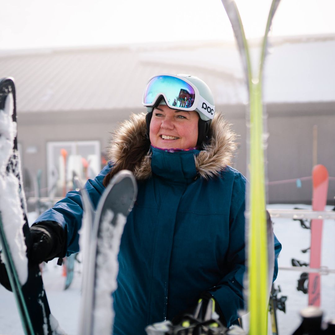 Smiling woman holding skis near a ski rack, wearing the Cartel Brooklyn Stretch Women's Plus Size Ski Jacket in teal.