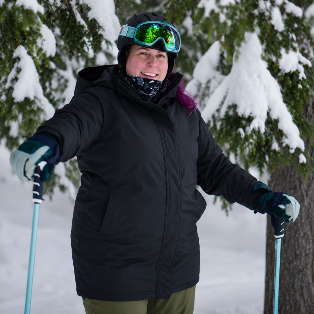 Smiling woman skiing on a snowy trail between evergreen trees wearing the black Brooky Long Stretch Jacket, ski helmet, and goggles.
