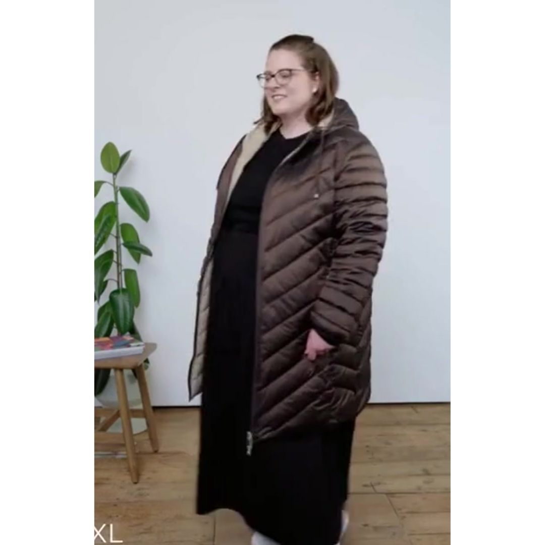 Plus size woman wearing a long brown puffer coat indoors with a plant and wooden stool in the background.