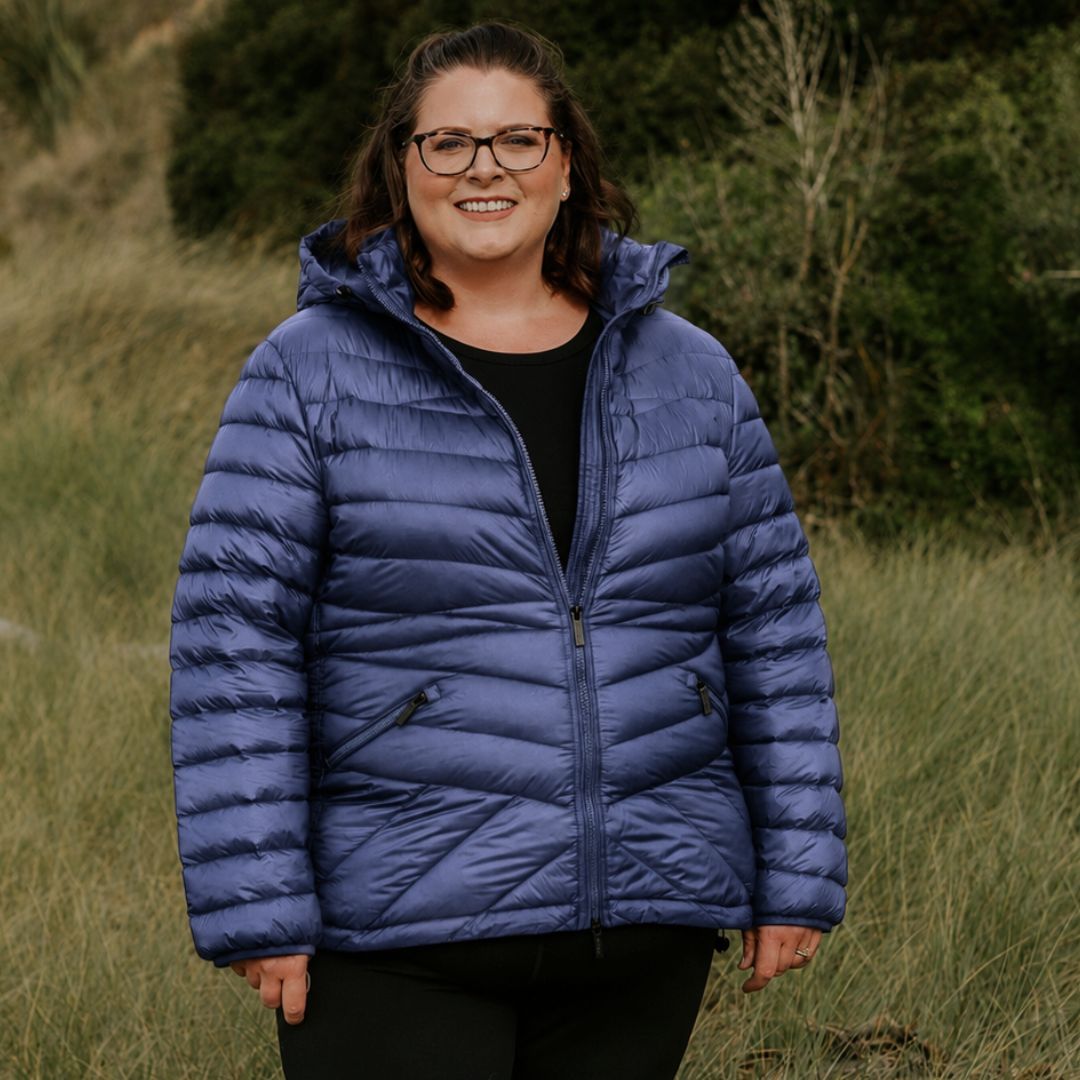 Front view of a smiling woman wearing the zipped moonlight blue Moke Cushla Women's Plus Size Down Jacket in a grassy field setting.