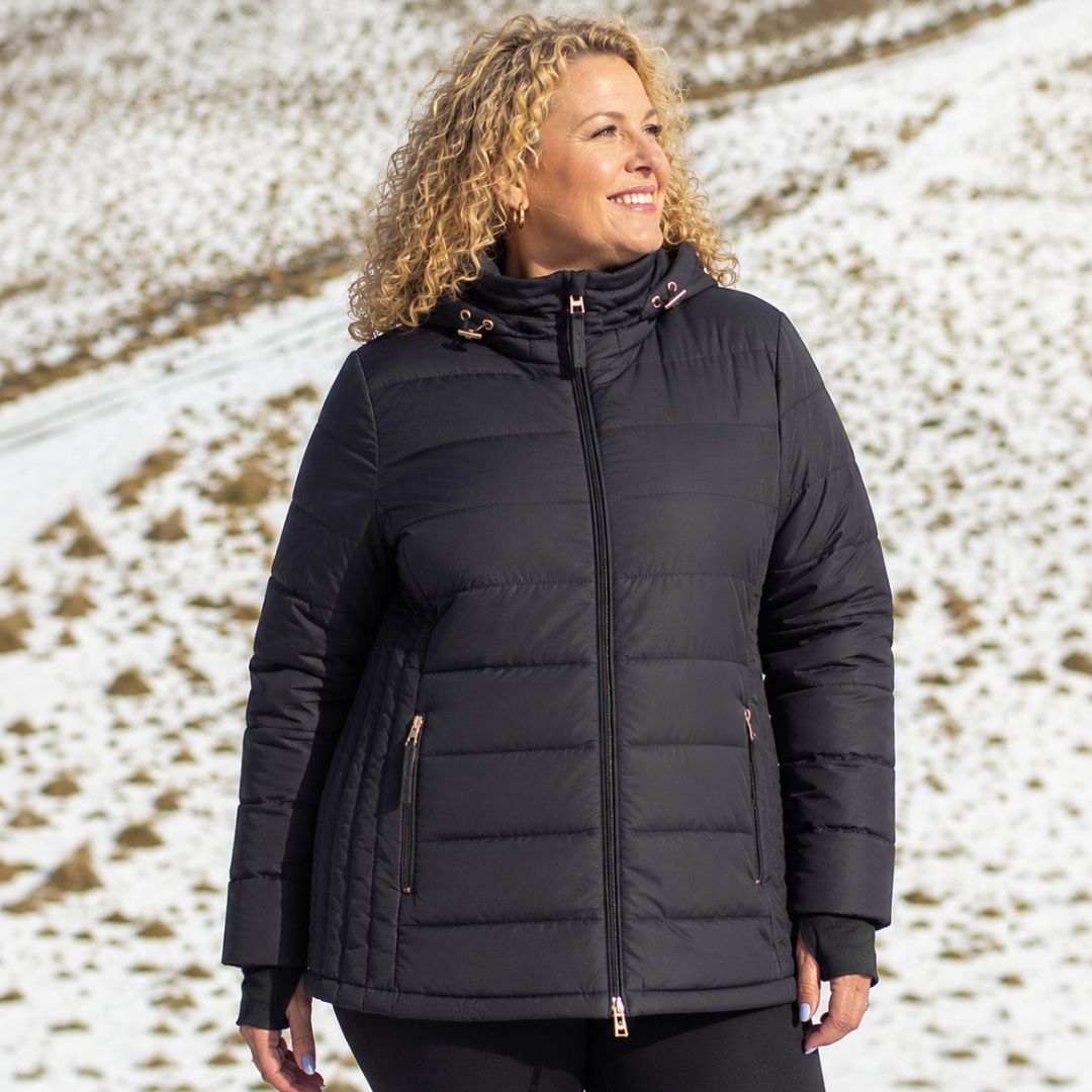 Smiling curly-haired woman standing in a snowy field wearing the zipped Moke Luciana Women's Down Puffer Jacket in black.