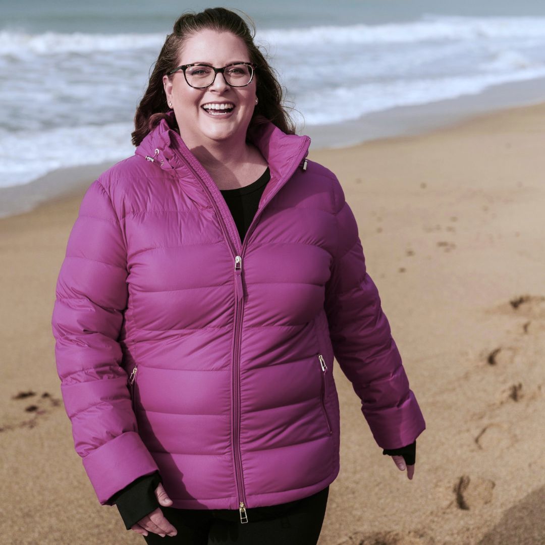 Laughing woman with glasses walking on the beach wearing the zipped Moke Luciana Women's Down Puffer Jacket in rose violet.