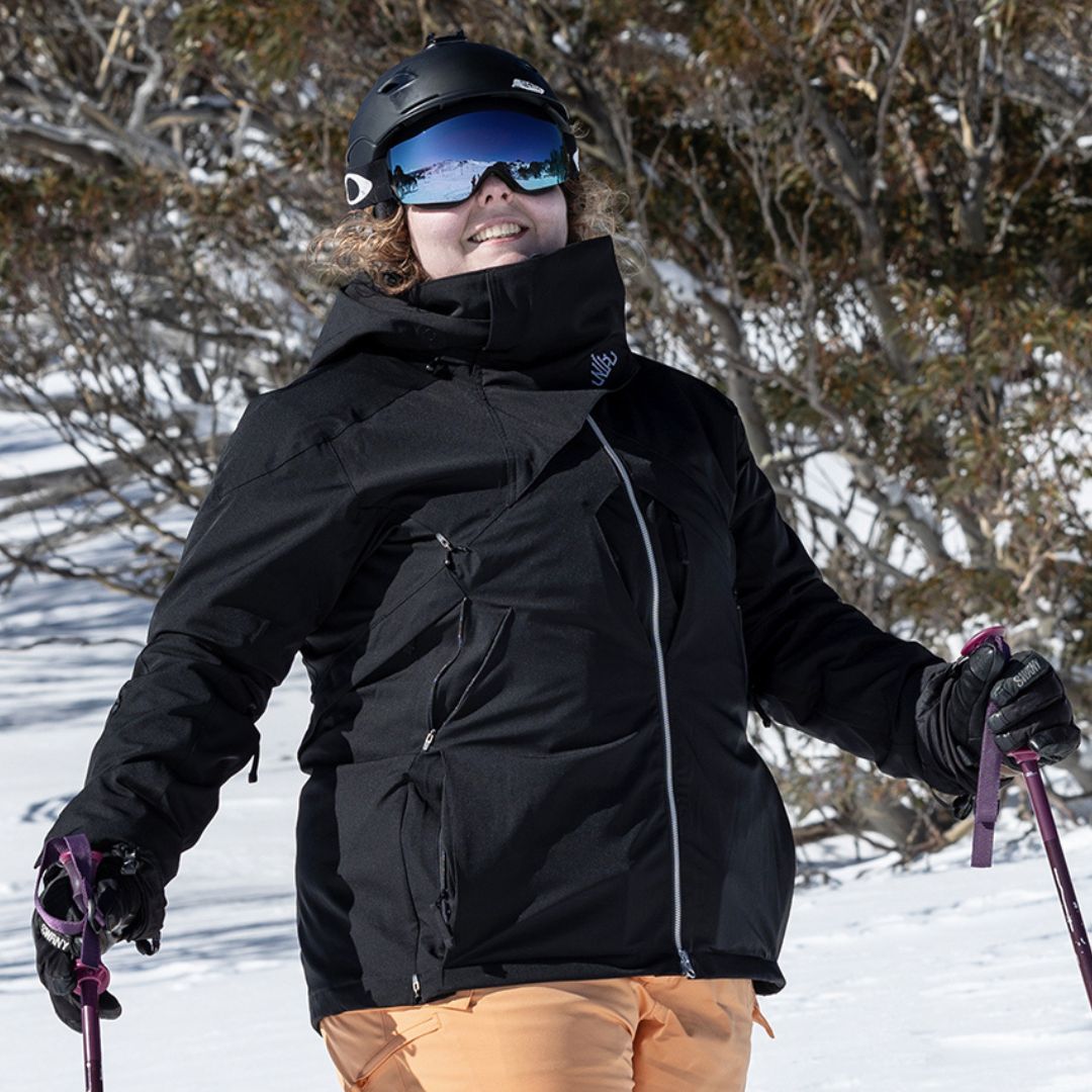 Person in black jacket and goggles standing on a snowy slope with trees in the background