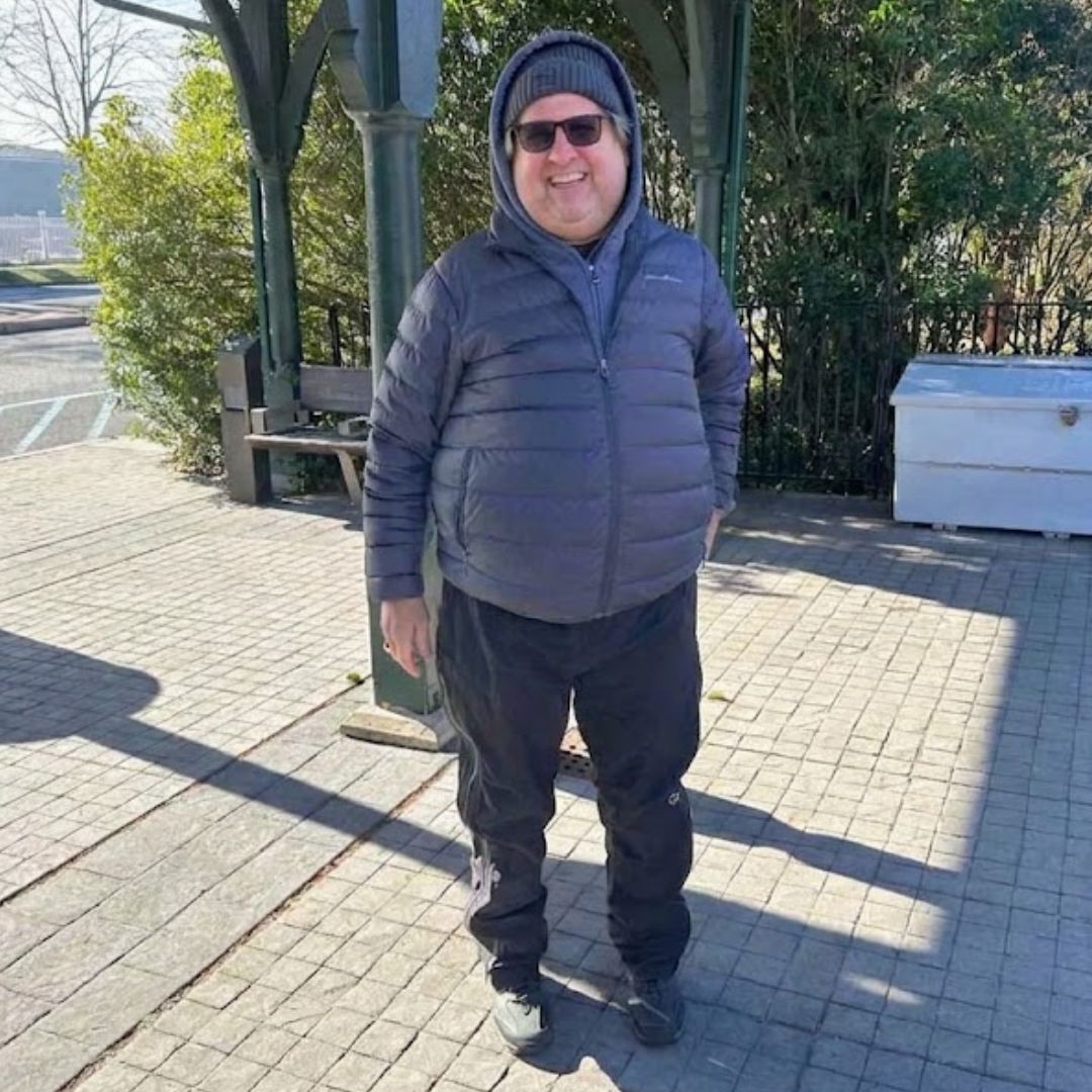 A plus size man wearing a dark gray puffer jacket, black sweatpants, and a gray beanie stands on a paved outdoor walkway under a green pavilion structure.