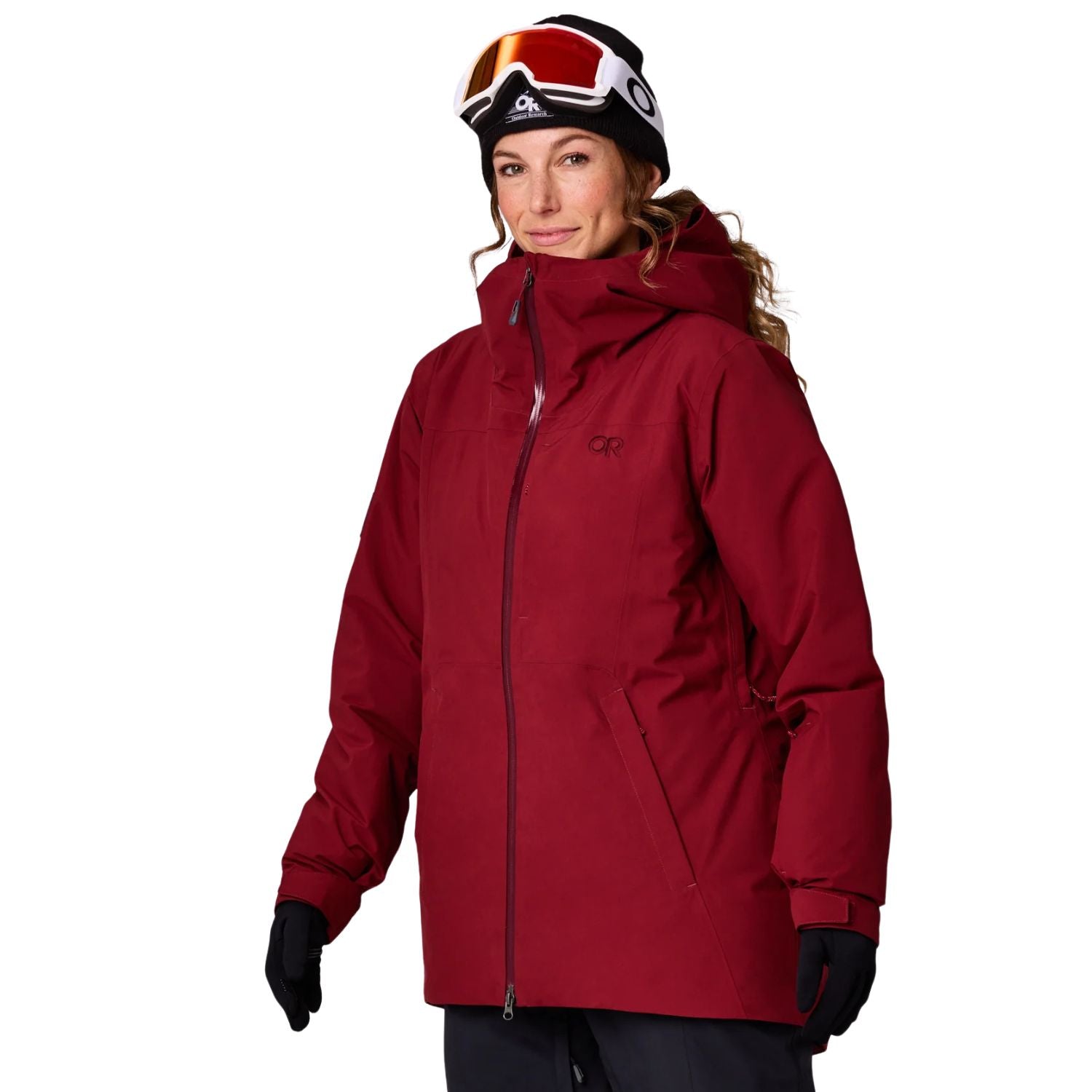 A plus size woman wearing a red hooded ski jacket, black gloves, a black beanie with ski goggles, and dark pants, photographed against a plain white studio background.