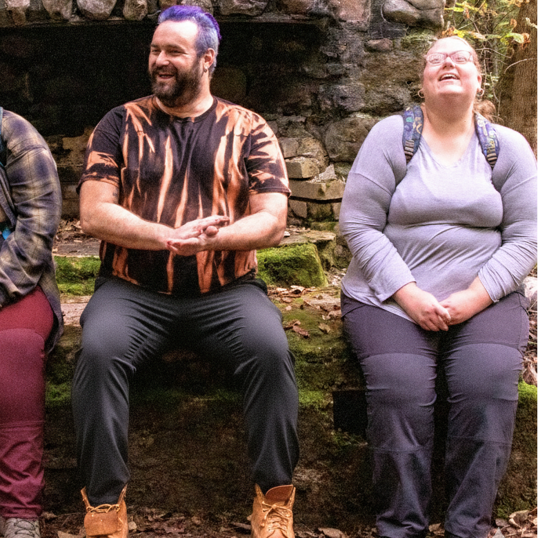 Three plus size people sitting in front of a stone wall in a forest setting