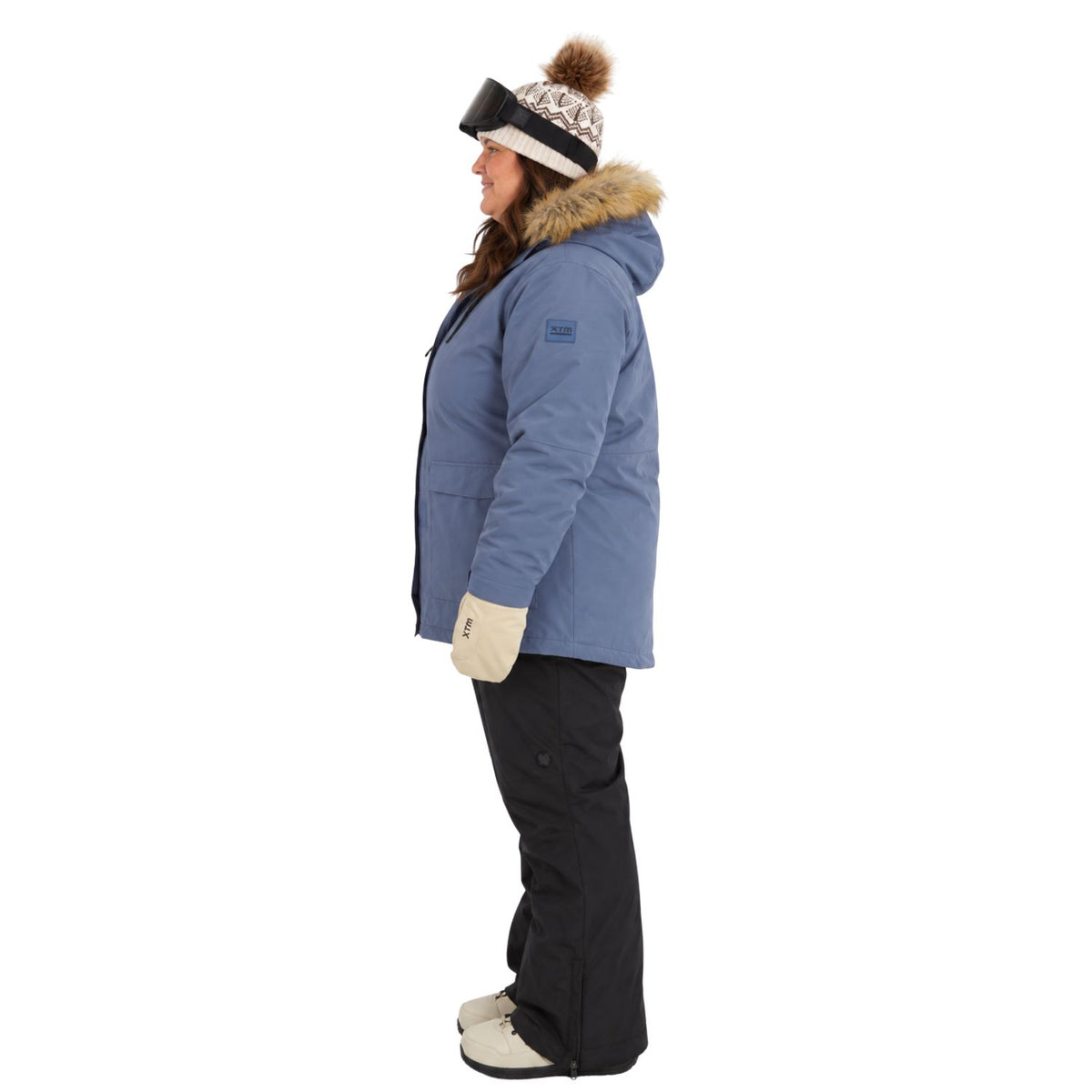 A woman shown in profile wearing a light blue winter parka with a brown faux-fur trimmed hood, black snow pants, cream gloves, and a patterned knit beanie with a pom-pom, against a plain white background.