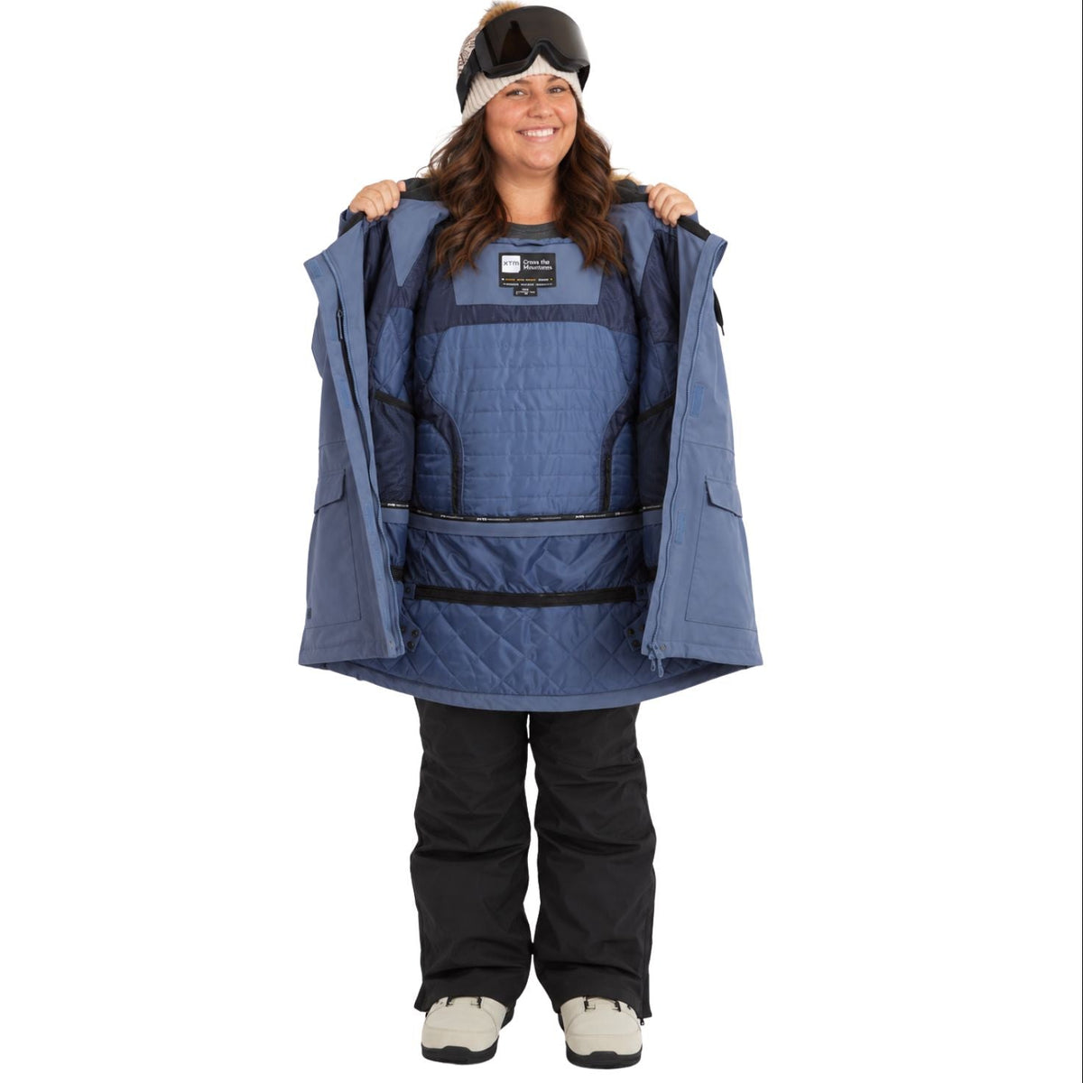 A woman wearing a blue quilted snowboard jacket with a built-in insulated liner, holding the jacket open to show the liner. She is also wearing black snow pants, a black and white beanie, and goggles, photographed against a solid white background.