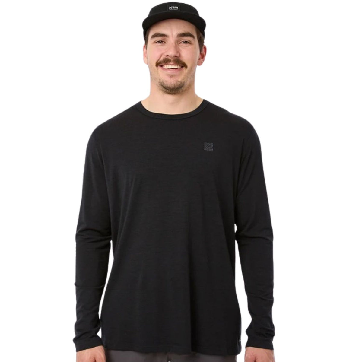 A man wearing a black long-sleeve crewneck shirt with a small tonal logo on the left chest, paired with a black cap, against a solid white background.