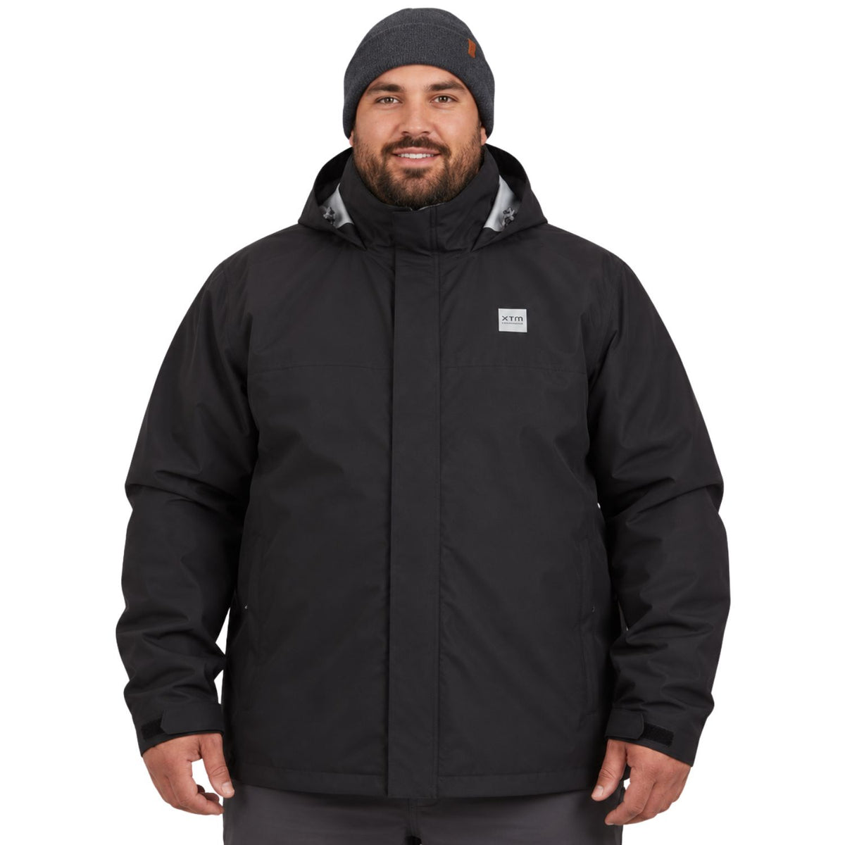 A big man wearing a black hooded rain jacket and a dark gray beanie, standing against a plain white background. The jacket has a small square logo patch on the left chest.