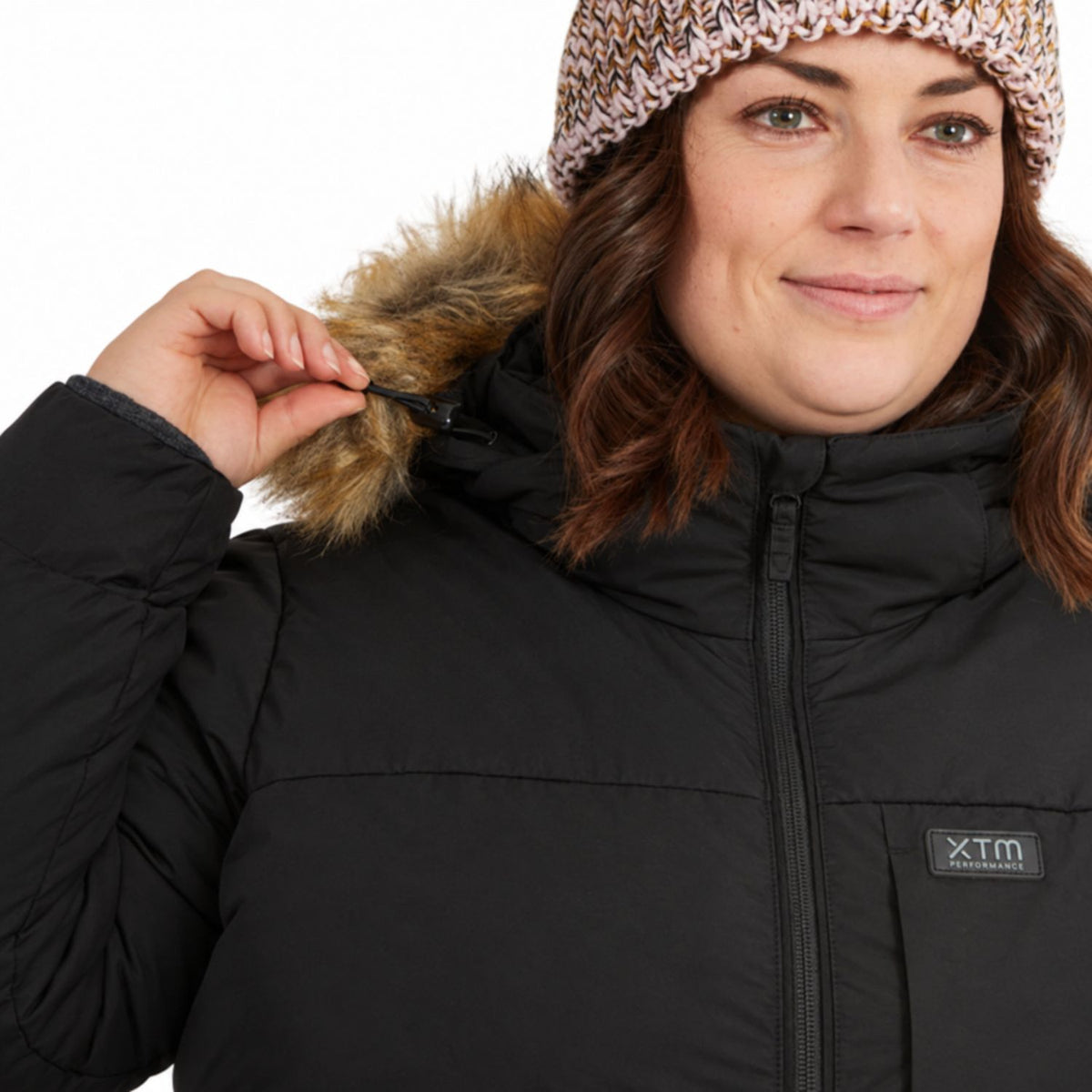 A close-up of a size 22 woman wearing a black winter puffer jacket with a brown faux-fur trimmed hood and a knit beanie. The jacket has a visible 'XTm' logo patch on the chest.